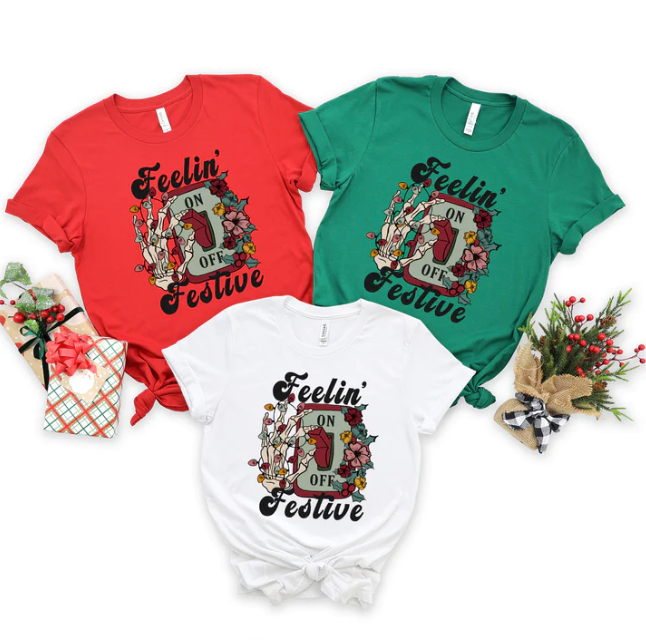 Three women's red, white, and green t-shirts feature a graphic design of a light switch and skeleton hand, wrapped in Christmas lights. The words "Feeling Festive" complete the design.